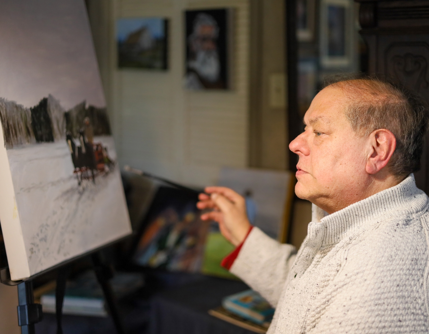 Photo of Chris Finkle on the right painting a landscape on a canvas on the left while wearing a light beige sweater. the background shows a home with many paintings