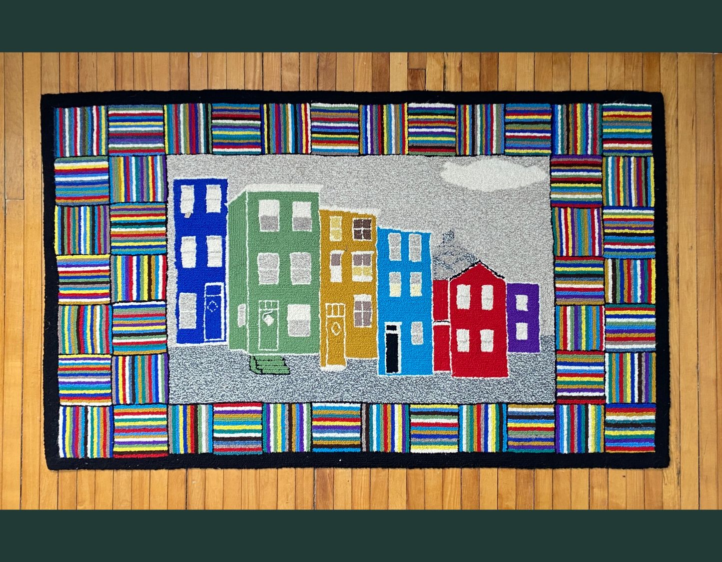 Image shows a wooden panel wall with a rug hung on the wall. The rug is a rectangle with colourful homes on a street. The colours are purple, red, light blue, yellow, light green and dark blue. The surrounding area is in the shape of squares with a series of colourful straight lines. The squares with straight lines alternate horizontal and vertical. The colours inlcude white, black, bright yellow, pink and purple. The background is dark teal.